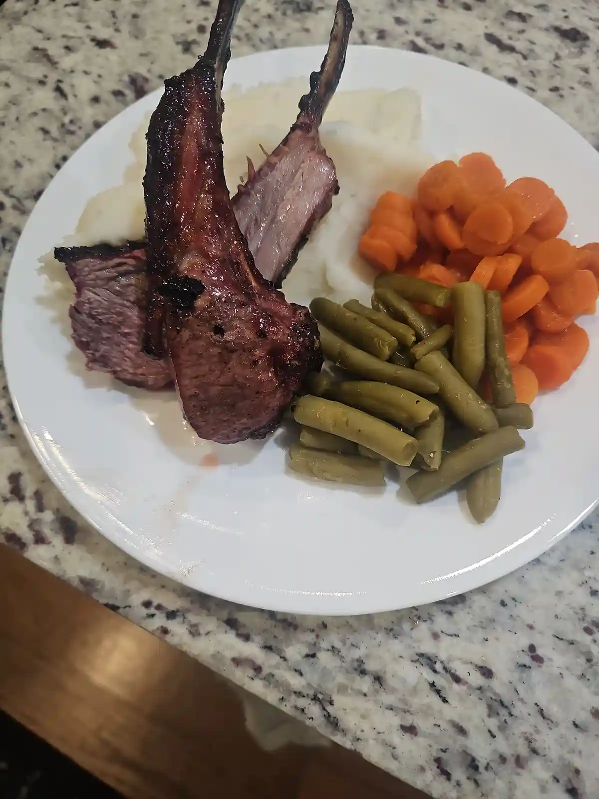 A white plate holds two grilled lamb chops standing upright on a bed of mashed potatoes, accompanied by a serving of sliced…