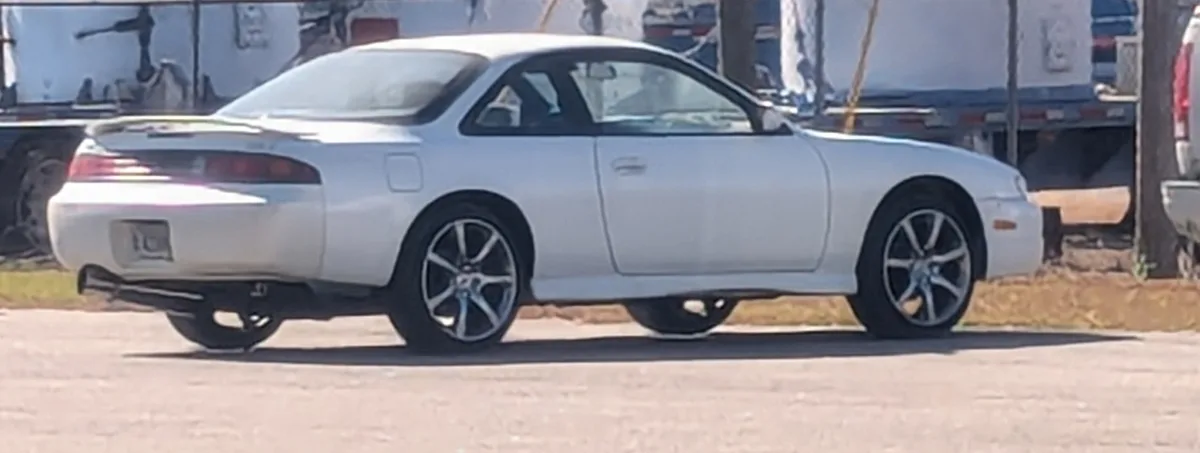 A white 1990s-era coupe, likely a Nissan 300ZX or similar, is parked on asphalt with a blurry background of trailers. The…