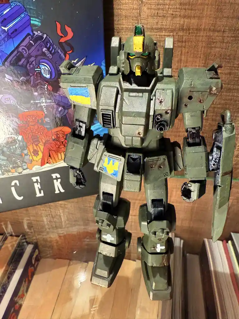 A heavily weathered, olive-green robot model stands upright on a wooden surface, its left shoulder and left thigh panels…