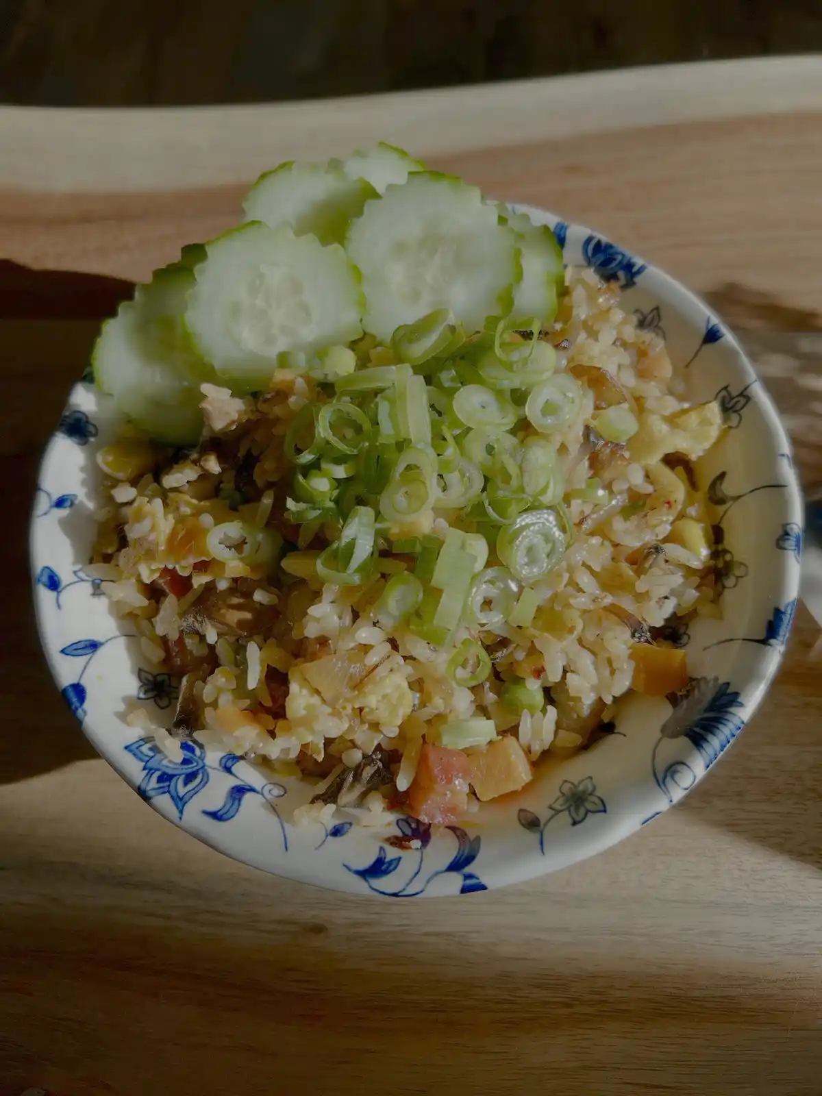 A close-up photo shows a bowl of fried rice topped with sliced cucumbers and chopped green onions, served in a blue and…