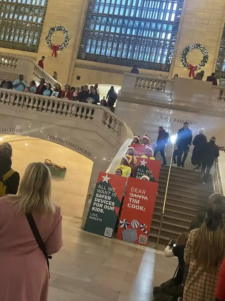 A group of people wearing Santa hats hold protest signs in a grand, festive public building with Christmas wreaths. One sign…