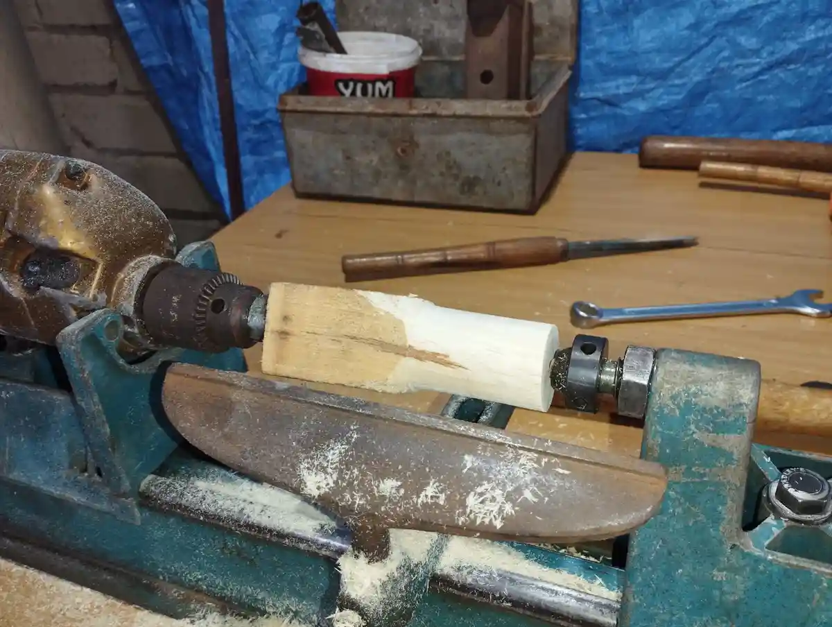 A close-up photo shows a wooden dowel being turned on a green metal lathe, with wood shavings scattered around. The…