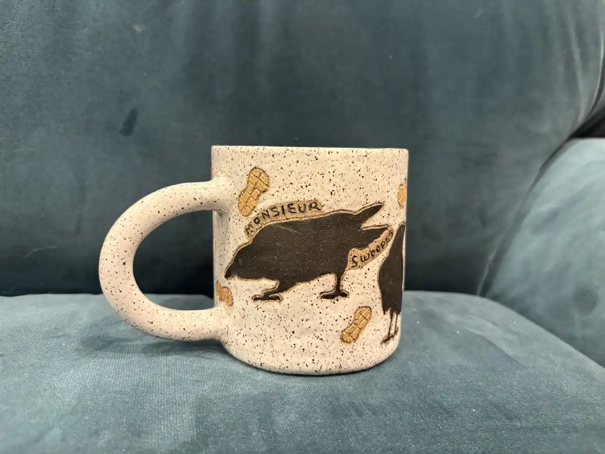 A speckled ceramic mug with raised black silhouettes of ravens and golden peanut illustrations sits on a teal fabric couch…