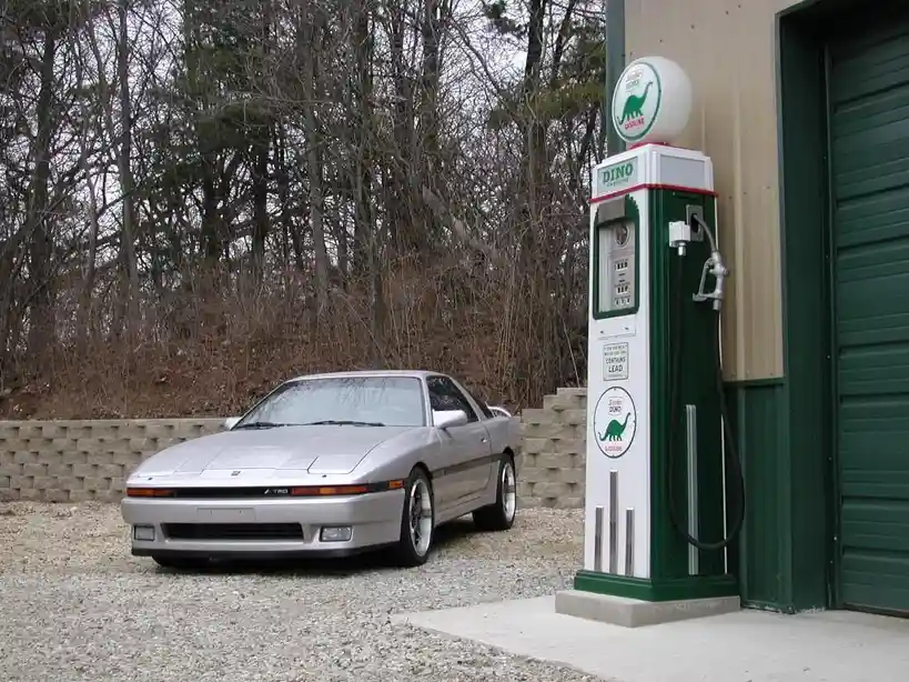 A silver Nissan 200SX sits parked on gravel next to a vintage green and white gas pump. The pump features a circular logo…
