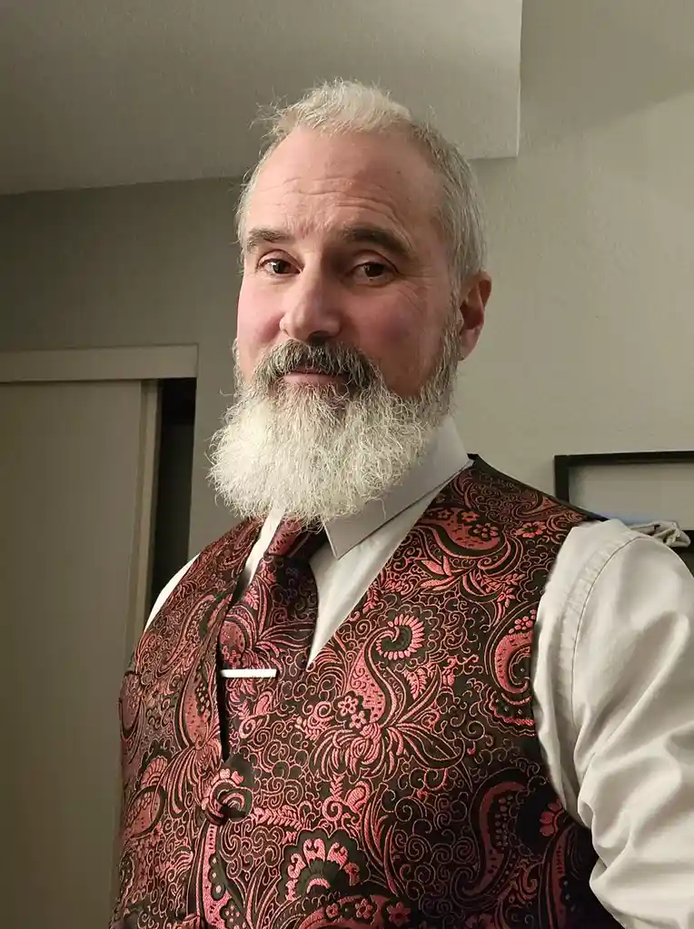 A middle-aged man with a full white beard and gray hair wears a white collared shirt, a red and black paisley vest, and a…