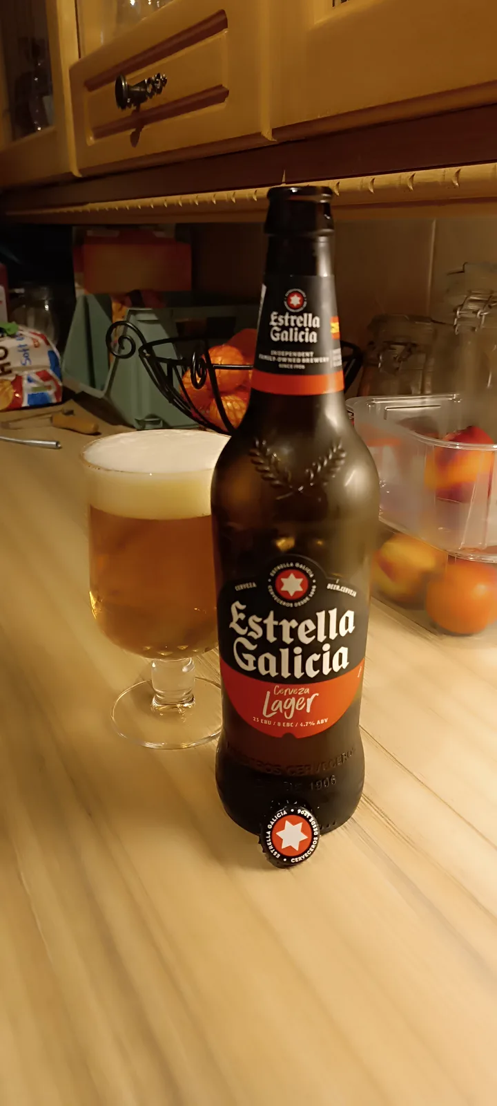 A bottle of Estrella Galicia beer sits on a wooden kitchen counter next to a glass filled with the beer and topped with…