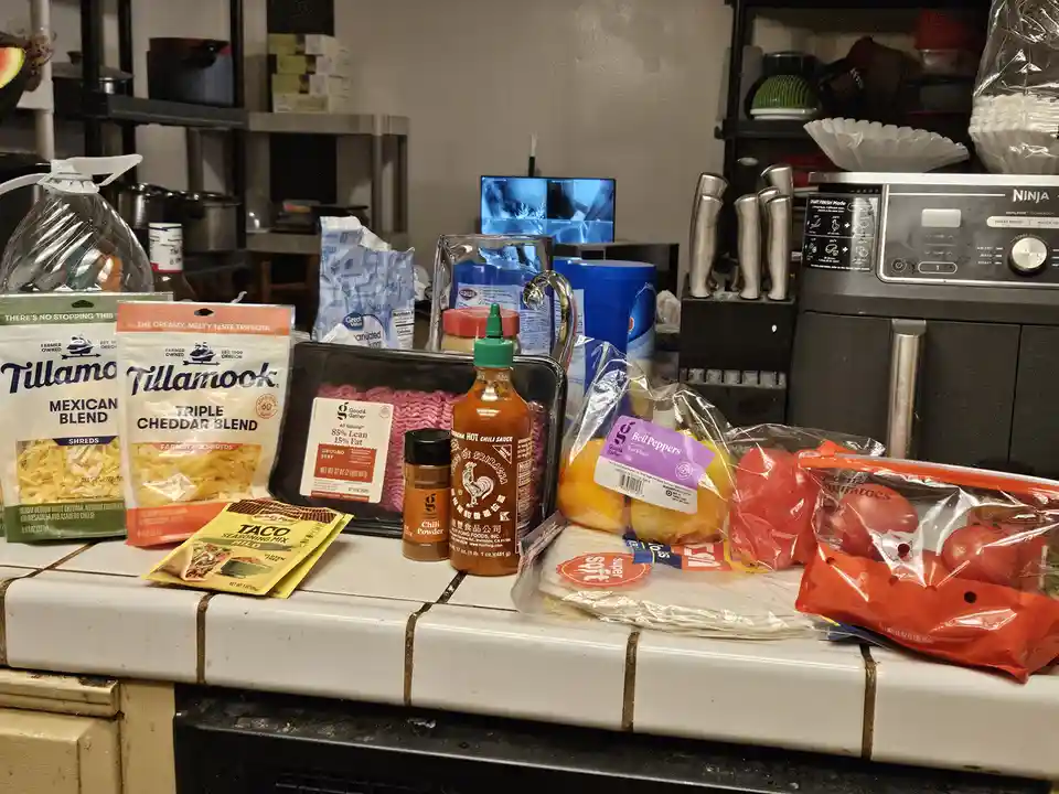 A cluttered kitchen counter is covered with ingredients for making tacos, including Tillamook cheese, ground beef, taco…