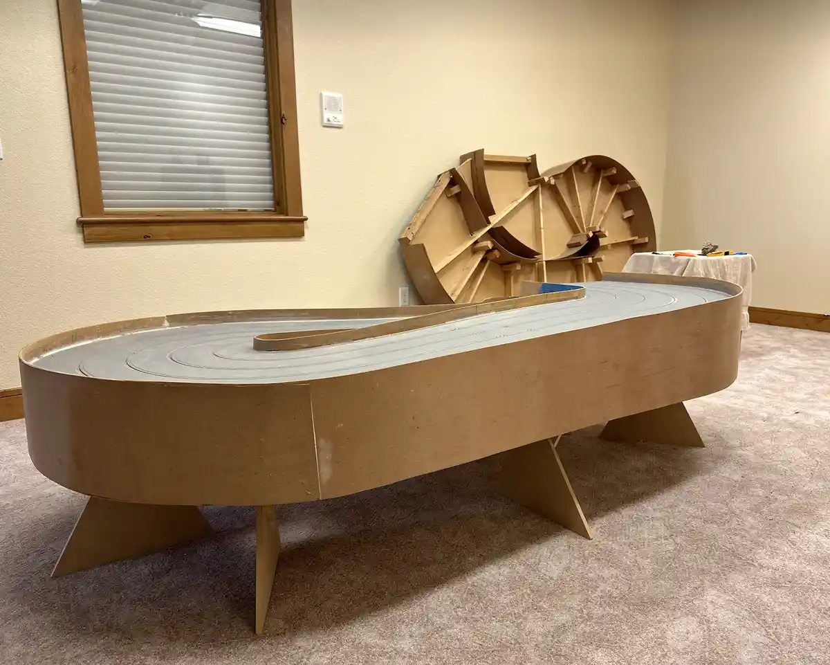 A large, custom-built race track made of cardboard sits on a carpeted floor in a room, featuring a curved grey track surface…