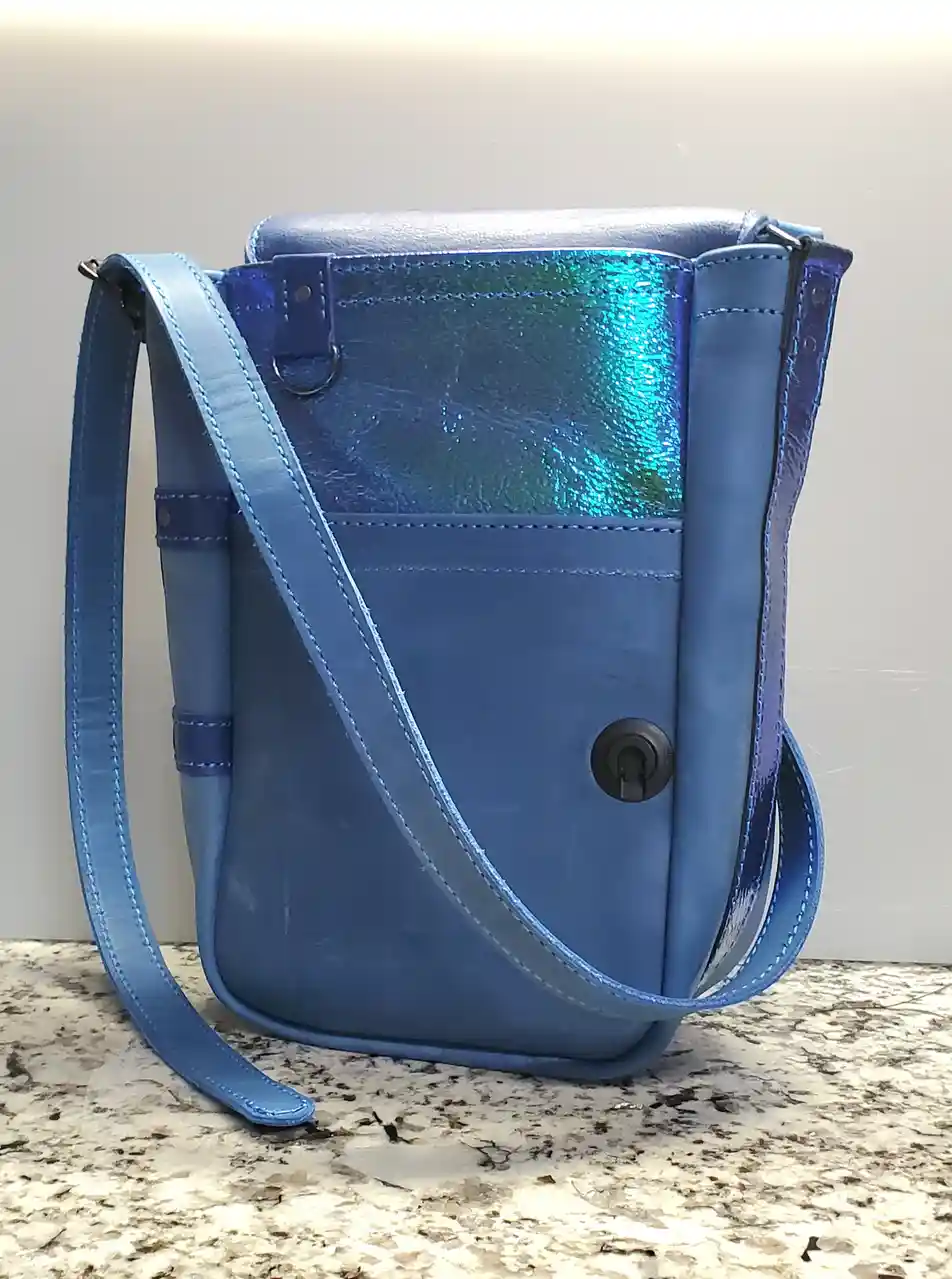 A blue leather crossbody bag with a shiny iridescent panel and a keyhole lock sits on a speckled countertop. The overlaid…
