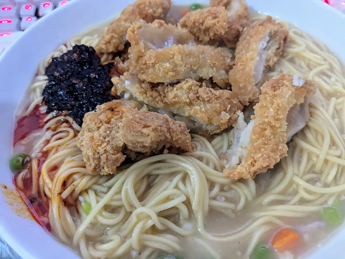 A close-up photo shows a white bowl filled with noodles, topped with crispy fried chicken pieces, a dollop of dark black…