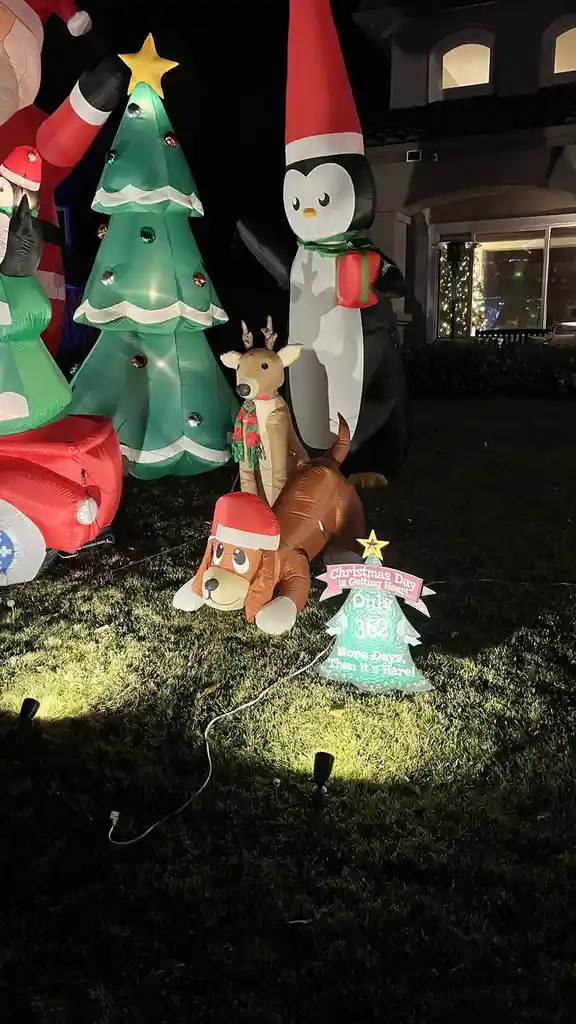 A nighttime photo of a front yard decorated with large, illuminated Christmas inflatable decorations including a Santa, a…