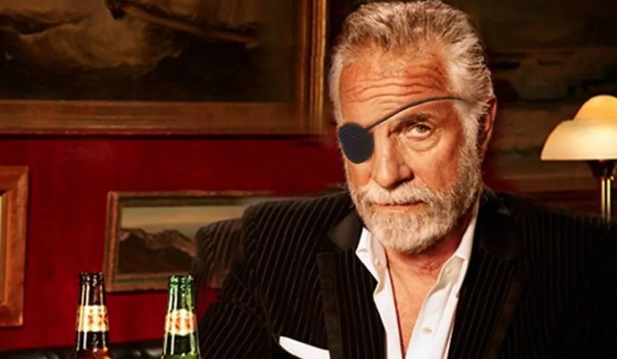 A man with a white beard and an eyepatch sits at a table with beer bottles, wearing a pinstripe suit and looking sternly at…