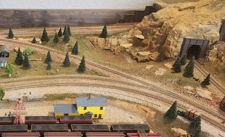 An overhead view of a detailed model train layout featuring miniature tracks, a yellow building, a rocky hill with a tunnel…