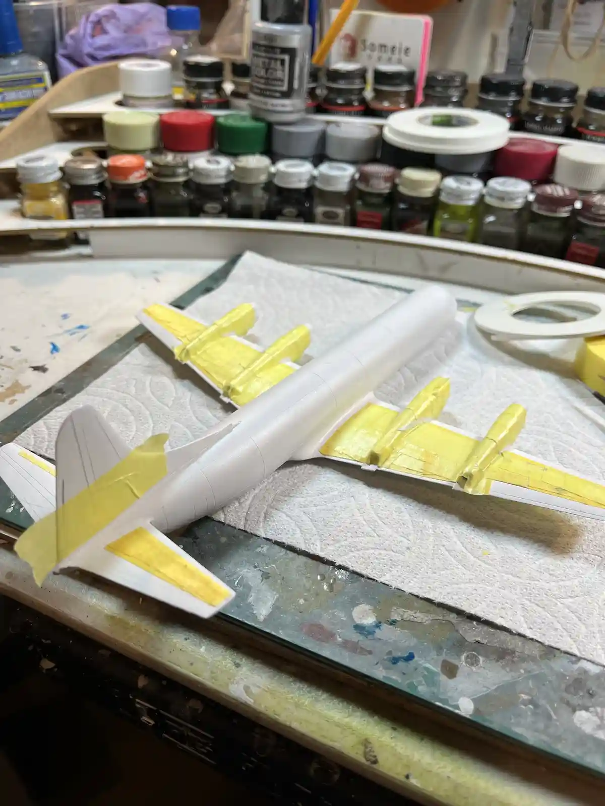 A white model airplane sits on a paper towel on a paint-splattered workbench, with yellow paint applied to its wings and…