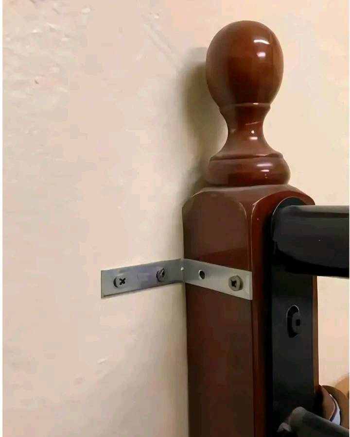 A close-up photo shows a shiny brown wooden stair railing post mounted to a wall with a simple metal bracket and screws. The…