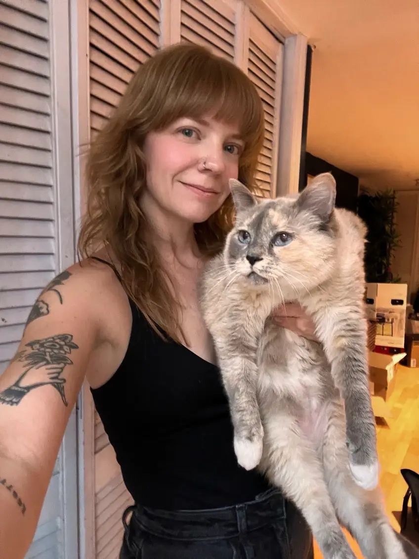 A woman with shoulder-length brown hair and tattoos on her left arm holds a fluffy, light-colored cat with blue eyes. She…