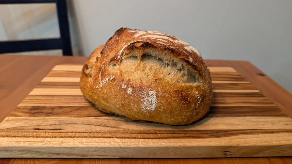 A freshly baked, rustic loaf of bread with a golden-brown crust and flour dusting sits on a striped wooden cutting board…
