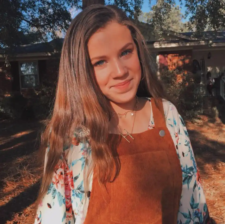 A young woman with long brown hair and blue eyes smiles slightly while wearing brown corduroy overalls over a floral shirt…
