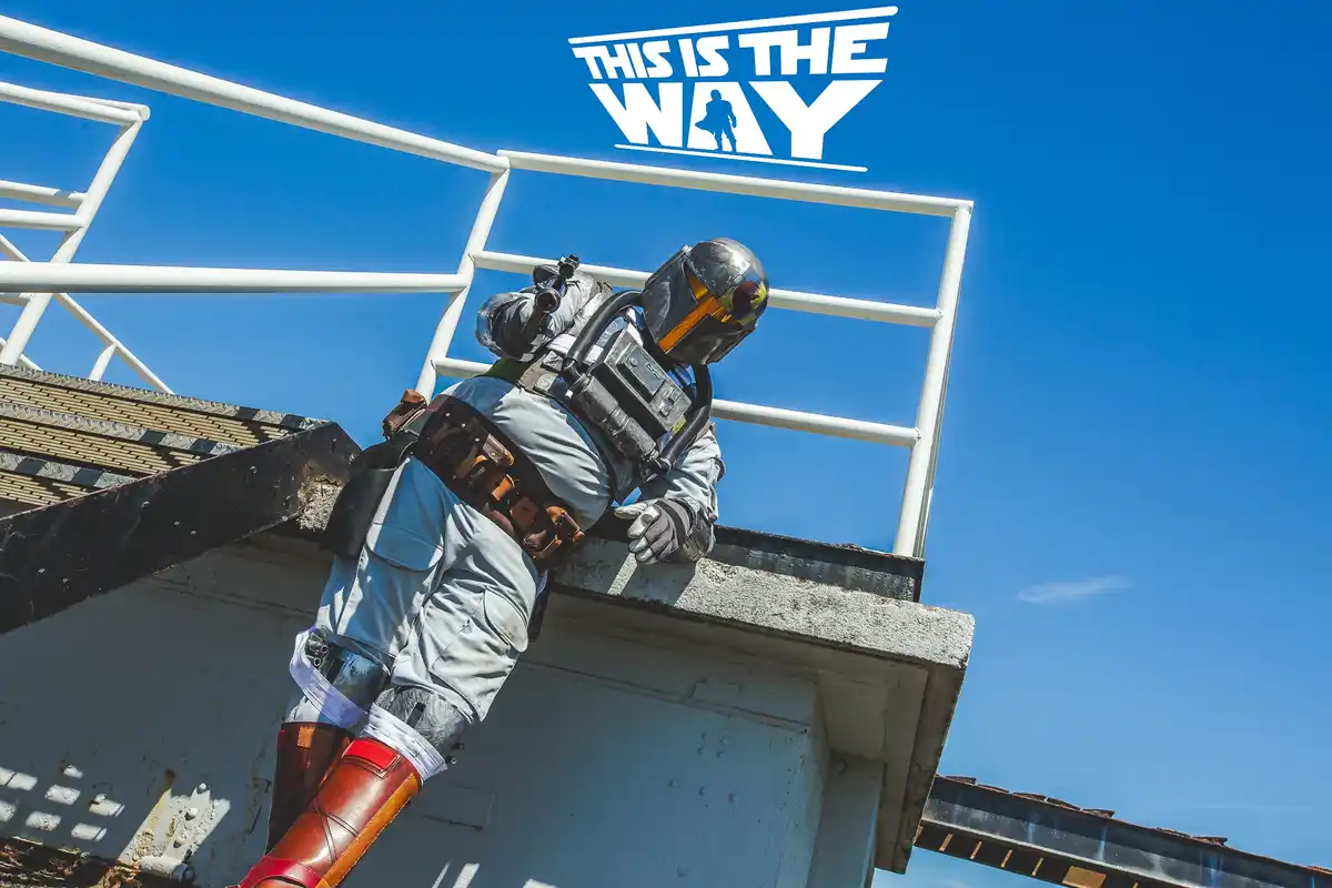 A person in a Mandalorian armor costume, holding a blaster, is leaning over the edge of a metal structure against a clear…