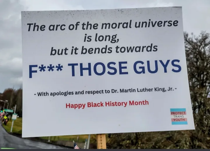 A white protest sign outdoors with black and red text humorously alters Dr. Martin Luther King Jr.'s famous quote to 'F***…