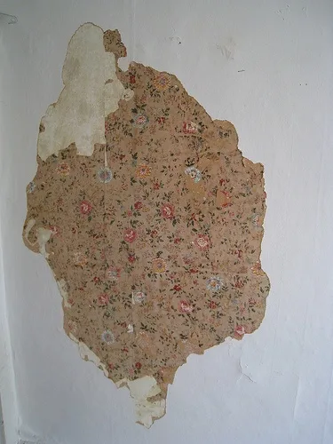 A large, irregularly shaped piece of floral wallpaper is peeling off a white wall, revealing the bare surface underneath…