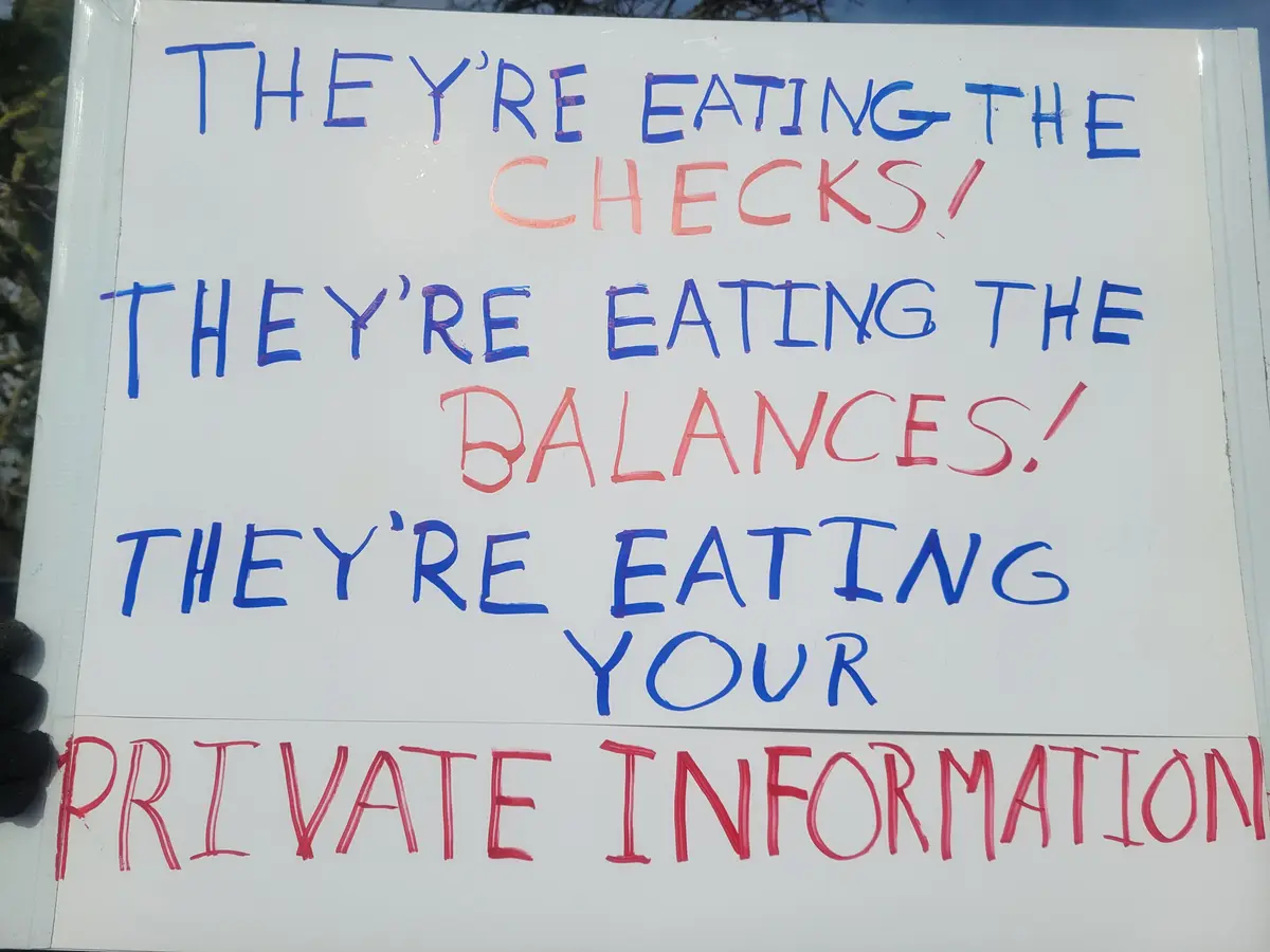 A white protest sign with handwritten text in blue and red marker, held by a gloved hand. The text reads: 'THEY'RE EATING…