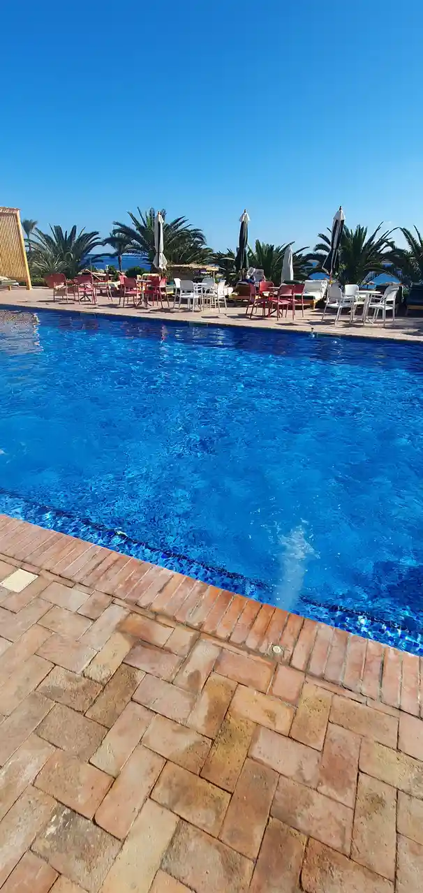 A bright, sunny photo of a large, clear blue swimming pool with a terracotta brick deck in the foreground. Around the pool's…