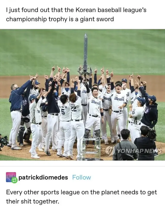 A baseball team in white uniforms celebrates on a field, raising their arms in triumph around a large, ornate sword standing…