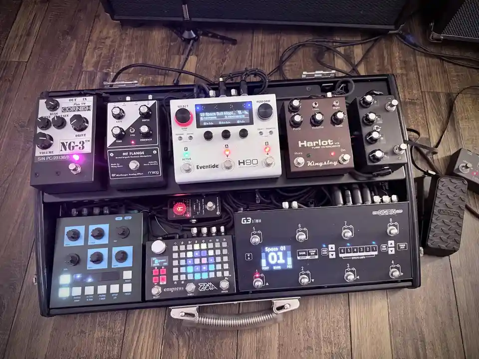 An overhead shot of a black pedalboard filled with numerous guitar effects pedals, including a Cornish NG-3, a Moog MF…