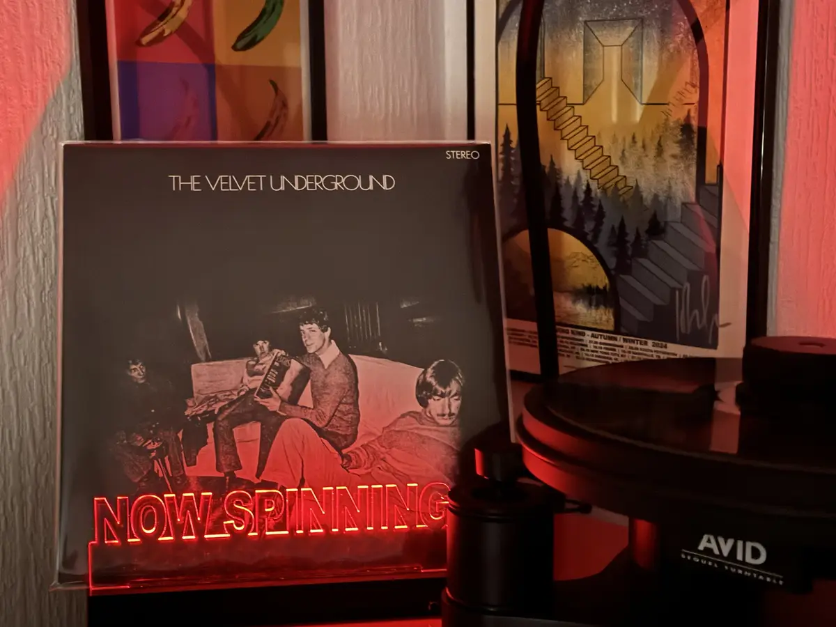 A vinyl record sleeve for 'The Velvet Underground' is propped up next to a turntable, with a glowing red neon sign that…