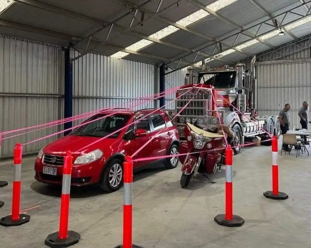 a red car, a red motorcycle, and a red semi-truck are parked inside a metal-sided warehouse, cordoned off by orange traffic…