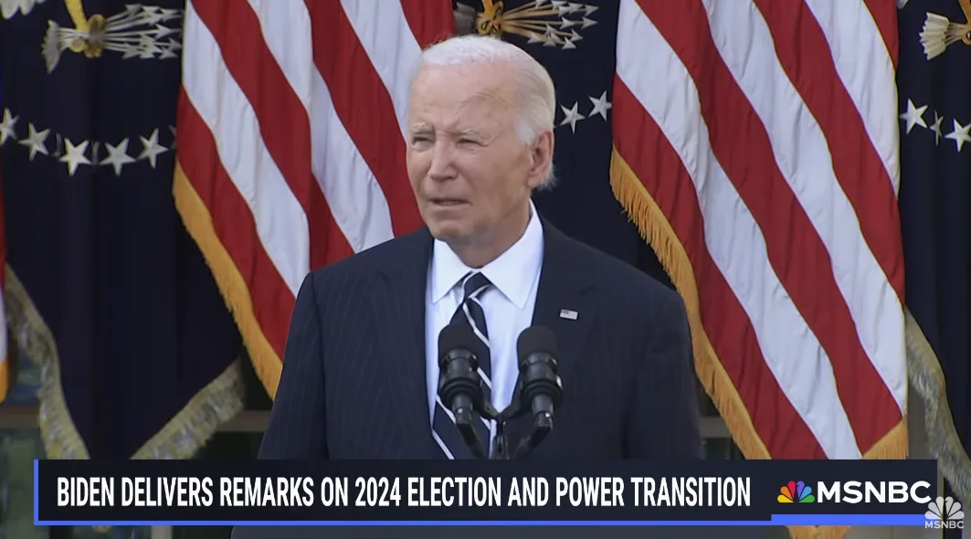 A man in a dark pinstripe suit and striped tie stands at a podium with two microphones, flanked by American flags. He has a…