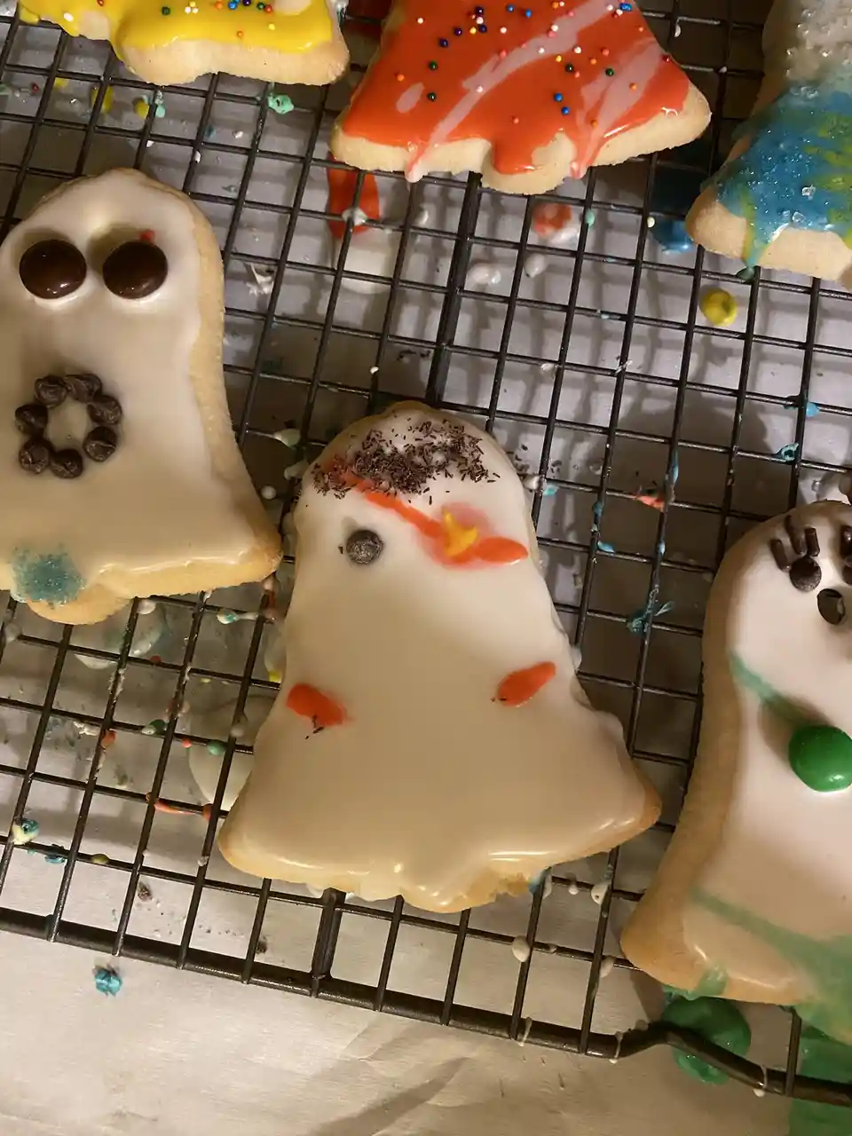 Several ghost-shaped sugar cookies with white icing, chocolate chip eyes, and messy decorations sit on a cooling rack. One…