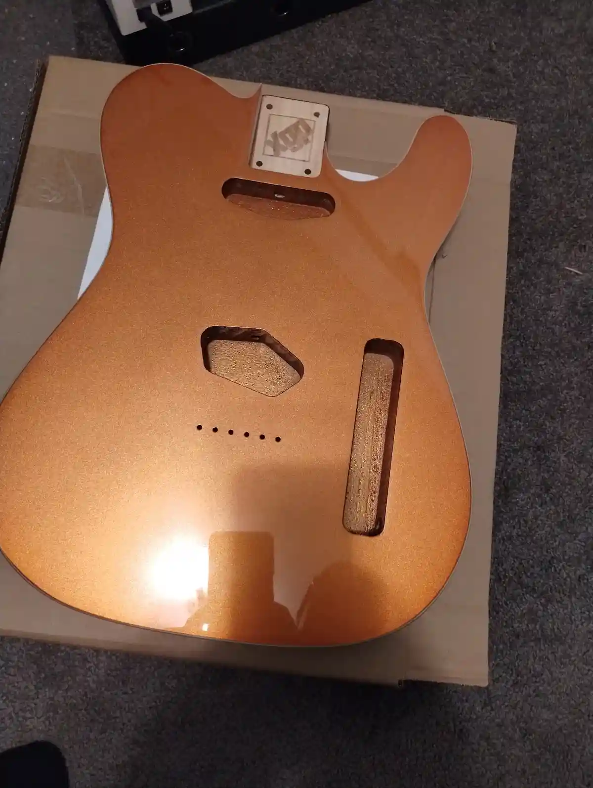 A close-up photo shows the back of a freshly painted, copper-colored electric guitar body resting on a cardboard box. The…