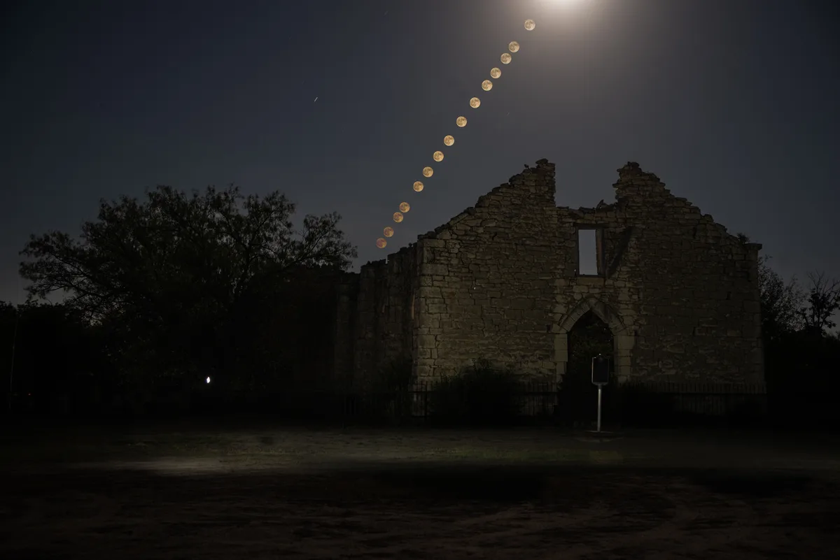 A night photograph shows the ruins of a stone building with a dark sky behind it, featuring a long diagonal trail of…
