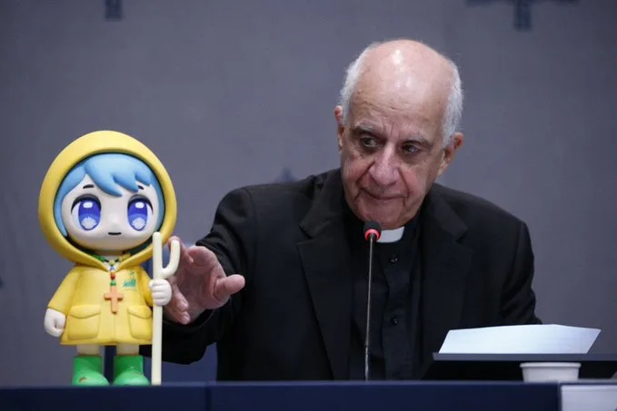 A man in a black clerical collar suit sits at a podium with a microphone, gesturing toward a yellow hooded mascot figure…