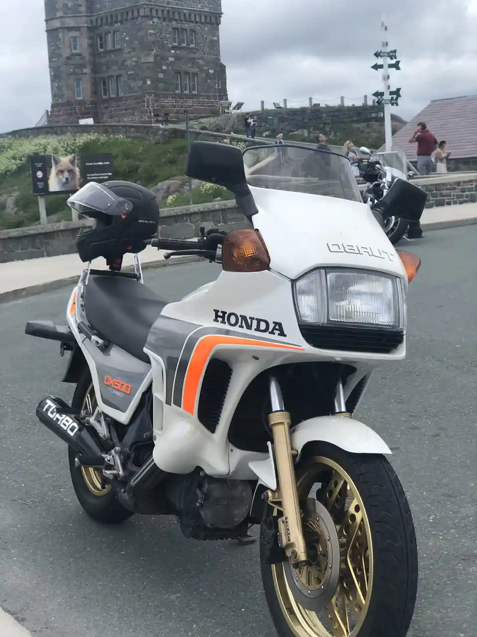 A white Honda CX500 Turbo motorcycle with gold wheels and orange accents is parked on a road near a stone tower. The word…