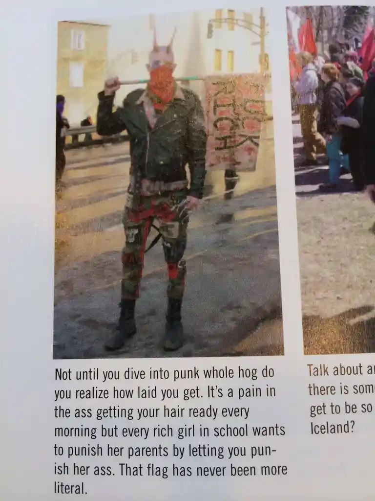 A person in punk attire, including a bandana, spiked hair, and camouflage pants, stands on a street holding a flag. The…