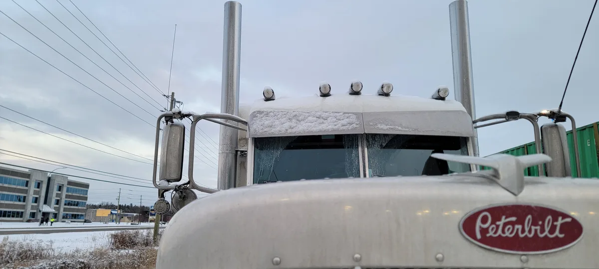 A close-up shot of a white Peterbilt semi-truck's cab, heavily covered in snow on its roof, hood, and windshield, with…