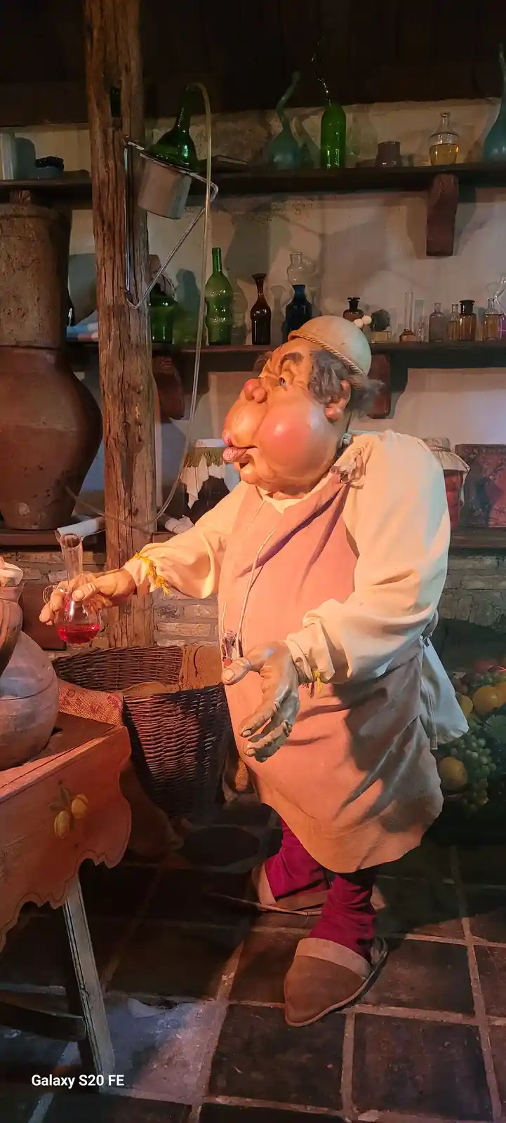 A grotesque, exaggerated wax figure of a plump, elderly man in a medieval-style lab coat and hat stands in a rustic…