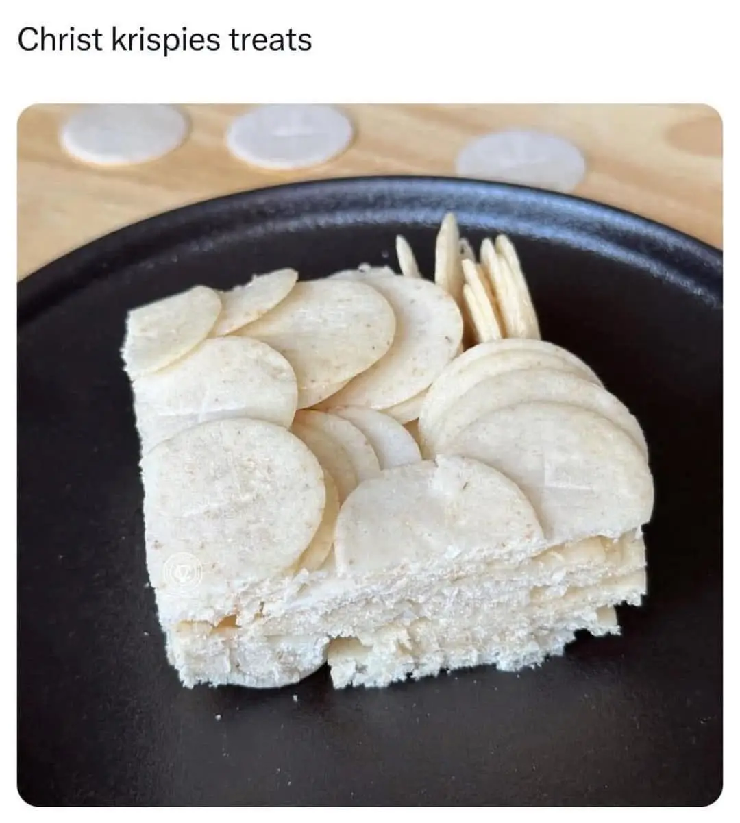 A close-up photo shows a square slice of a light-colored, layered dessert on a black plate, decorated with round, flat, pale…
