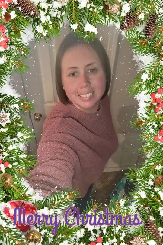 A selfie of a smiling woman in a pink sweater, framed by a festive holiday border with pine branches, pinecones, red…