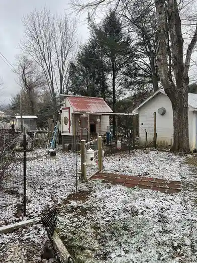 A snowy backyard scene shows a small white chicken coop with a red roof, surrounded by a wire fence and bare trees. A light…