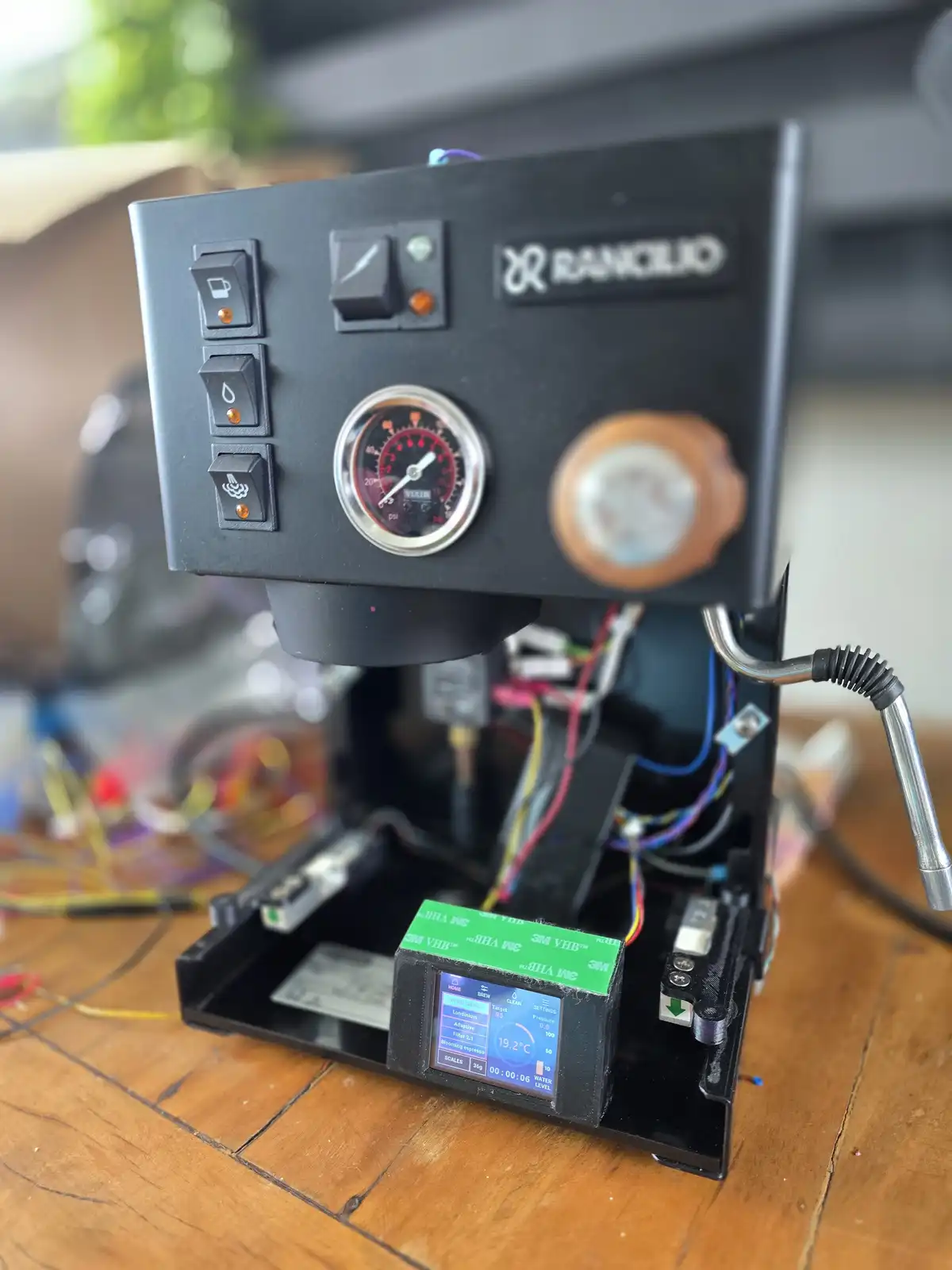 A black Rancilio espresso machine is shown with its internal wiring exposed and a digital display showing brewing parameters…