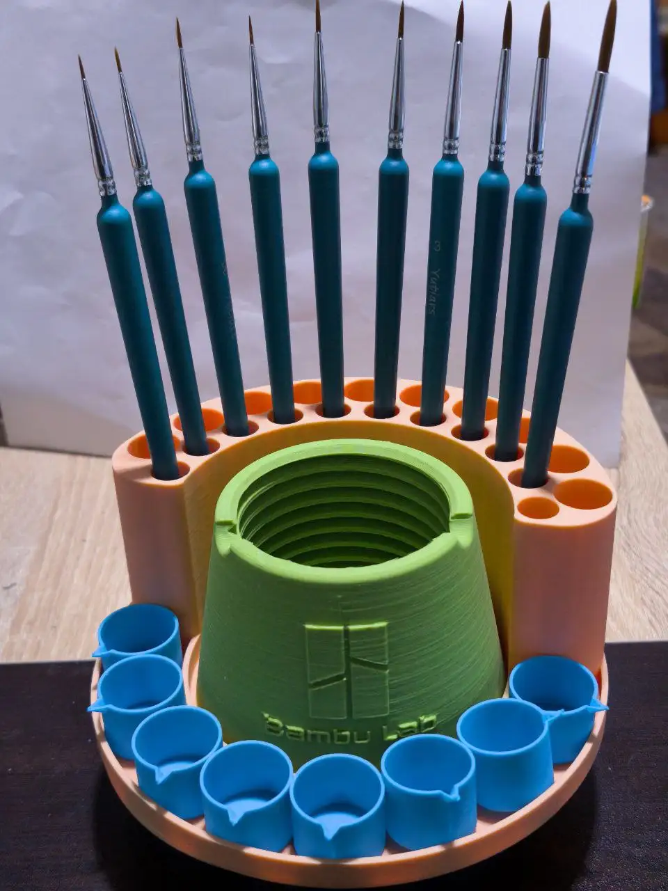 A 3D-printed orange and green stand holds multiple teal paintbrushes upright in circular slots, with a central green cup and…