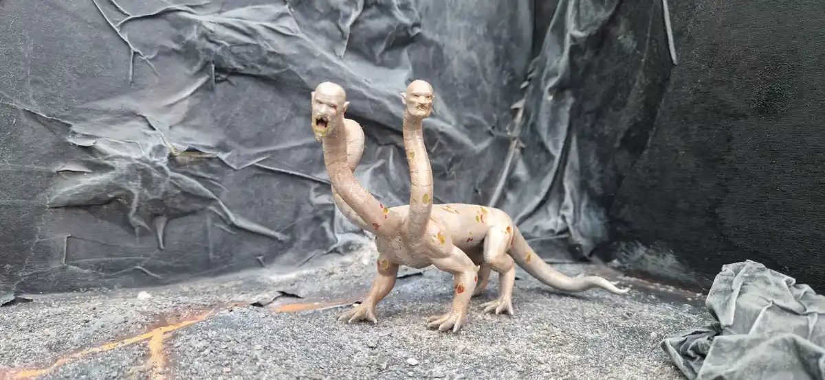 A pale, bipedal creature with two human-like heads stands on a gravel surface against a dark, crumpled backdrop. The…