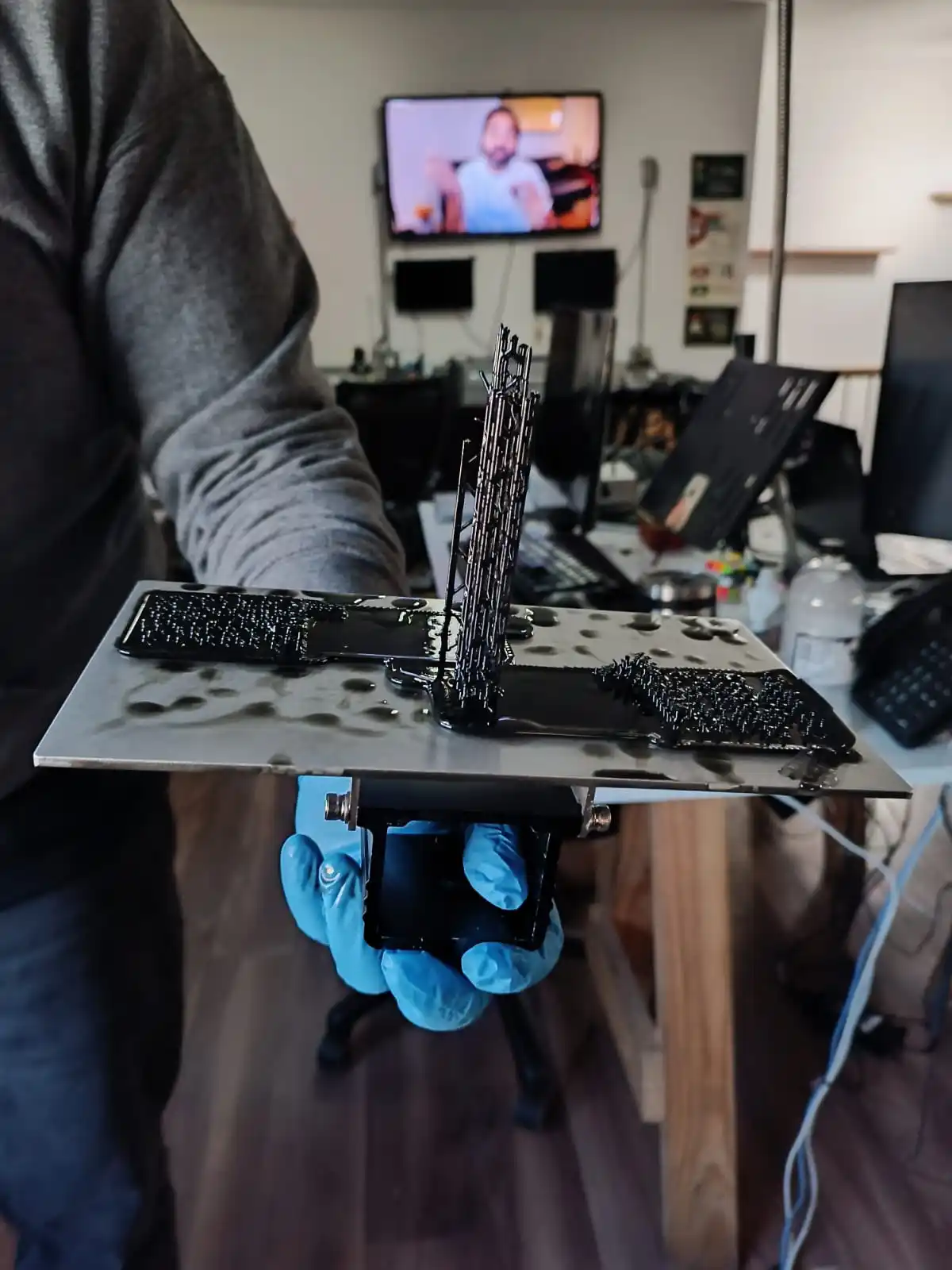 A person wearing blue gloves holds up a metal plate with a melted keyboard and its keys fused into a black, gooey, and…