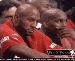 A sequence of six frames shows two bald men in red Miami Heat shirts sitting courtside during a basketball game. The man on…