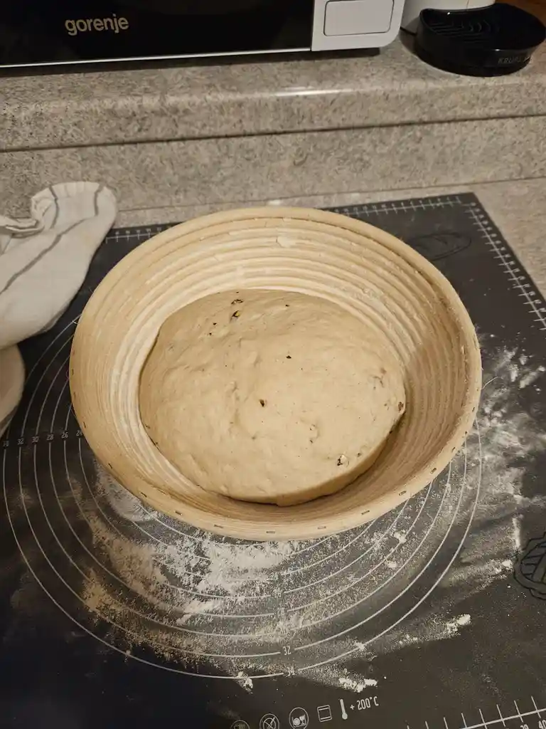 A round ball of risen bread dough sits in a wooden proofing basket on a black silicone baking mat dusted with flour. The mat…