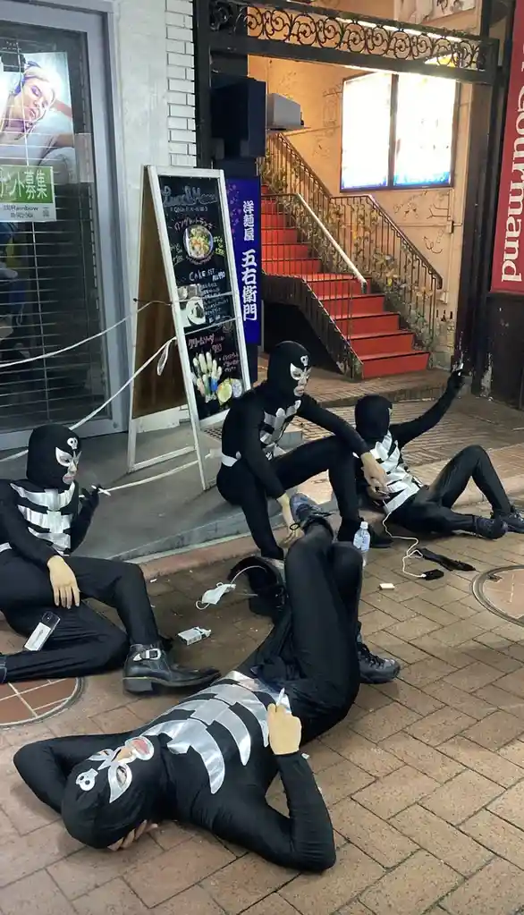 Four people dressed in black bodysuits with silver skeleton designs and skull masks are lounging casually on a city sidewalk…